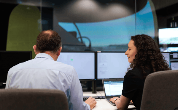 Two people sat at a desk in front of two big computer screens and a large window. The person on the right also has a laptop in front of them and is looking at the person on their left.